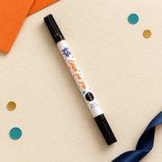 Scoff Paper edible ink pen resting on cream paper with orange envelope and navy ribbon